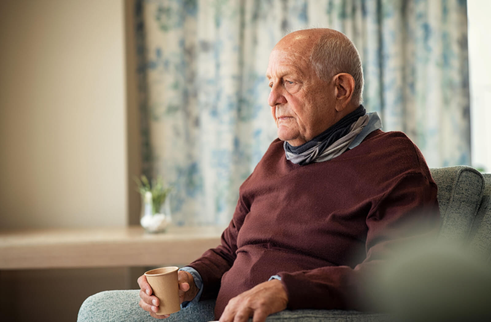 An older gentleman seated on a sofa, holding a paper cup, feeling lonely in his home.