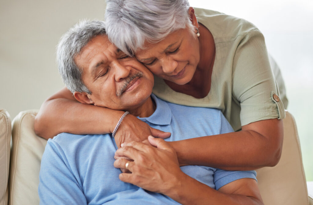 A senior woman comforting her senior husband who is sitting on a couch.