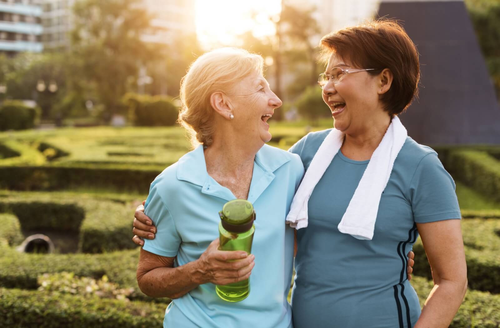 A pair of older adults stands arm in arm outside, laughing, after having finished an outdoor walk together