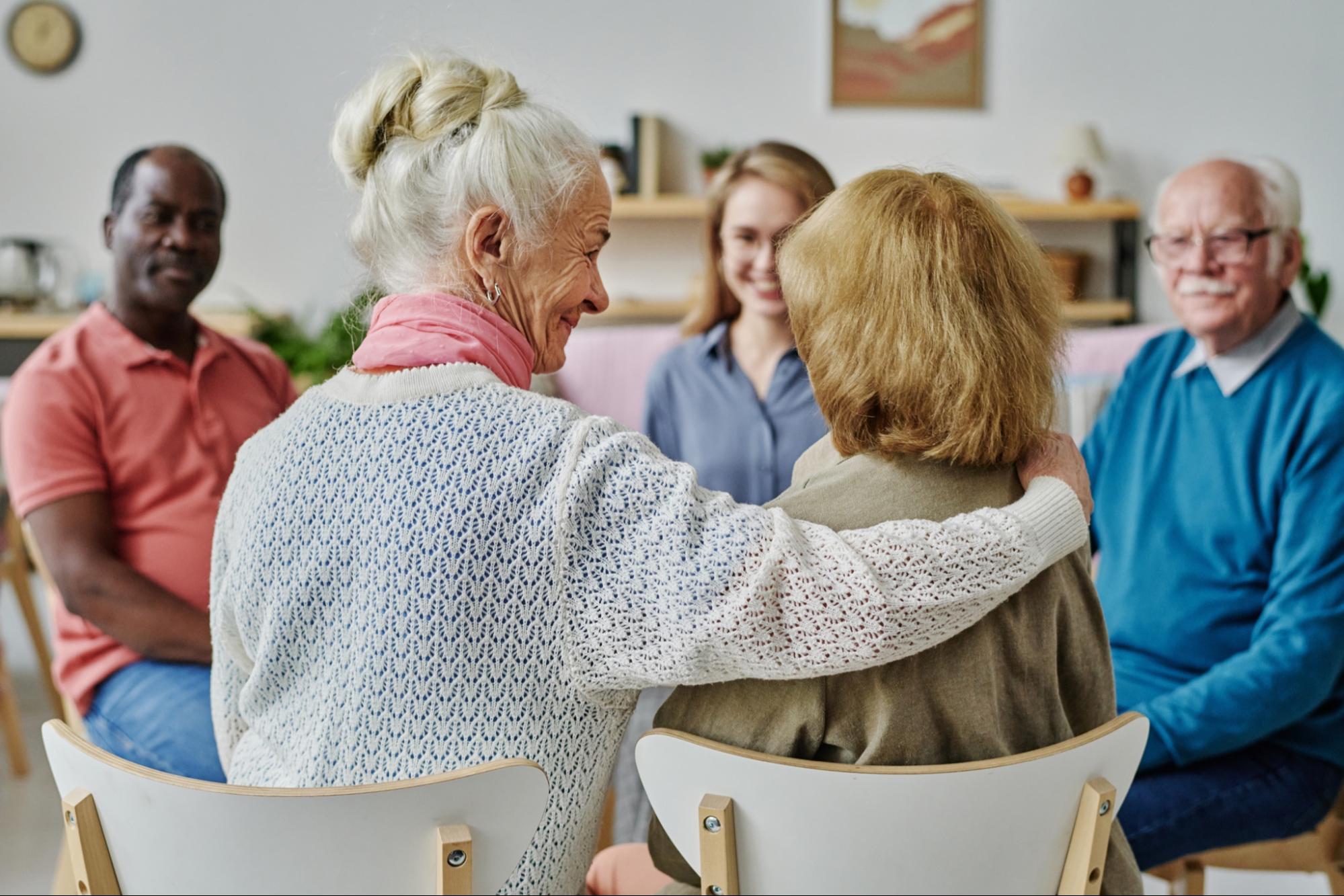 A group of older adults sitting in a circle, attending a support group in residence. The two women in front are embracing.